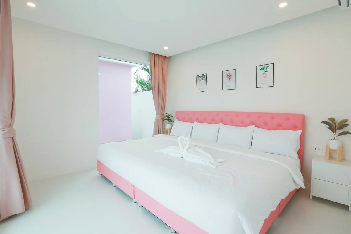 Un lit bien fait, avec du linge de lit blanc et une tête de lit rose, une table de chevet ornée d’une plante et des tableaux encadrés accrochés au mur.