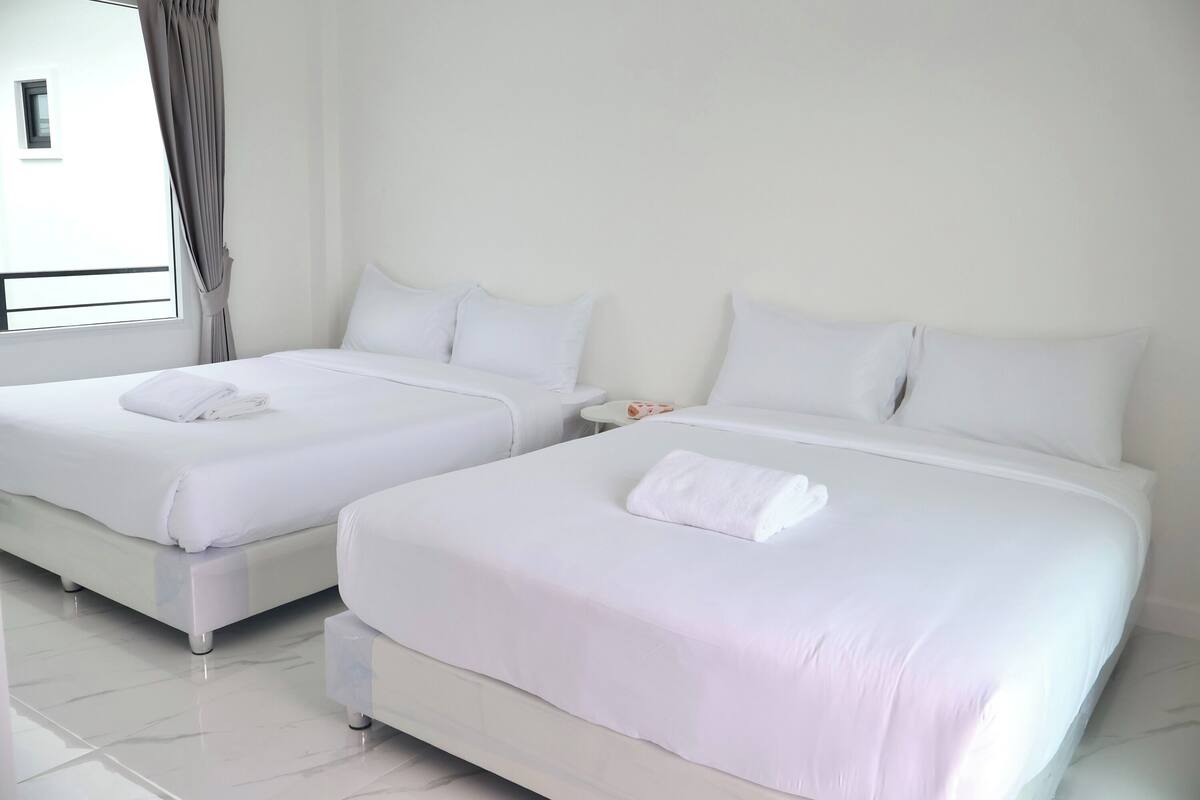 Deux lits bien faits, recouverts de draps blancs et agrémentés d’oreillers, une fenêtre avec des rideaux et un sol carrelé, propre et blanc.