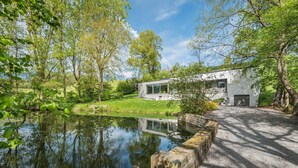 Une maison moderne à la façade en verre, entourée d’une végétation luxuriante et bordée d’un étang.