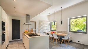 Une cuisine moderne dotée d’un îlot central, d’un coin repas avec un banc recouvert de tissu blanc et de grandes fenêtres offrant une vue sur la verdure.