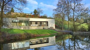Une maison moderne dotée d’une façade en verre, d’un bassin aux reflets changeants et d’un espace verdoyant.