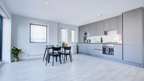 A modern kitchen with grey cabinets, a microwave, and an oven. There is a dining table with chairs and a potted plant. - Hybrid Resi - Uxbridge | 3 Bed Apartment (Uxbridge)