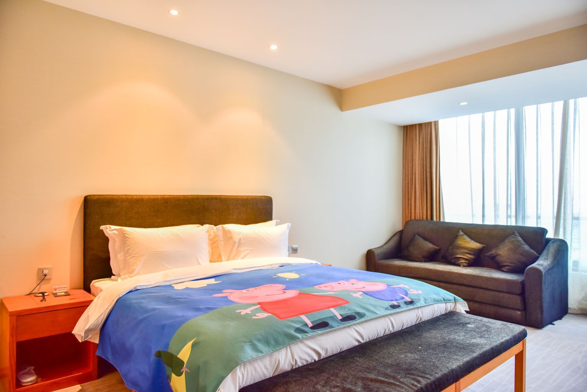 A hotel room with a bed, a bedside table, a bench, a sofa, and a large window with curtains.
