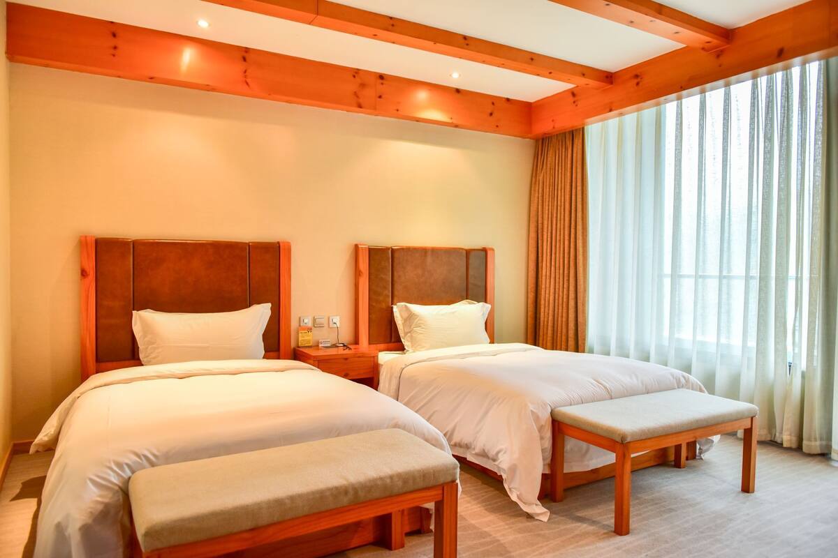 Habitación de hotel con dos camas, cabeceros de madera, un banco, una mesita de noche con lámpara y un ventanal con cortinas.
