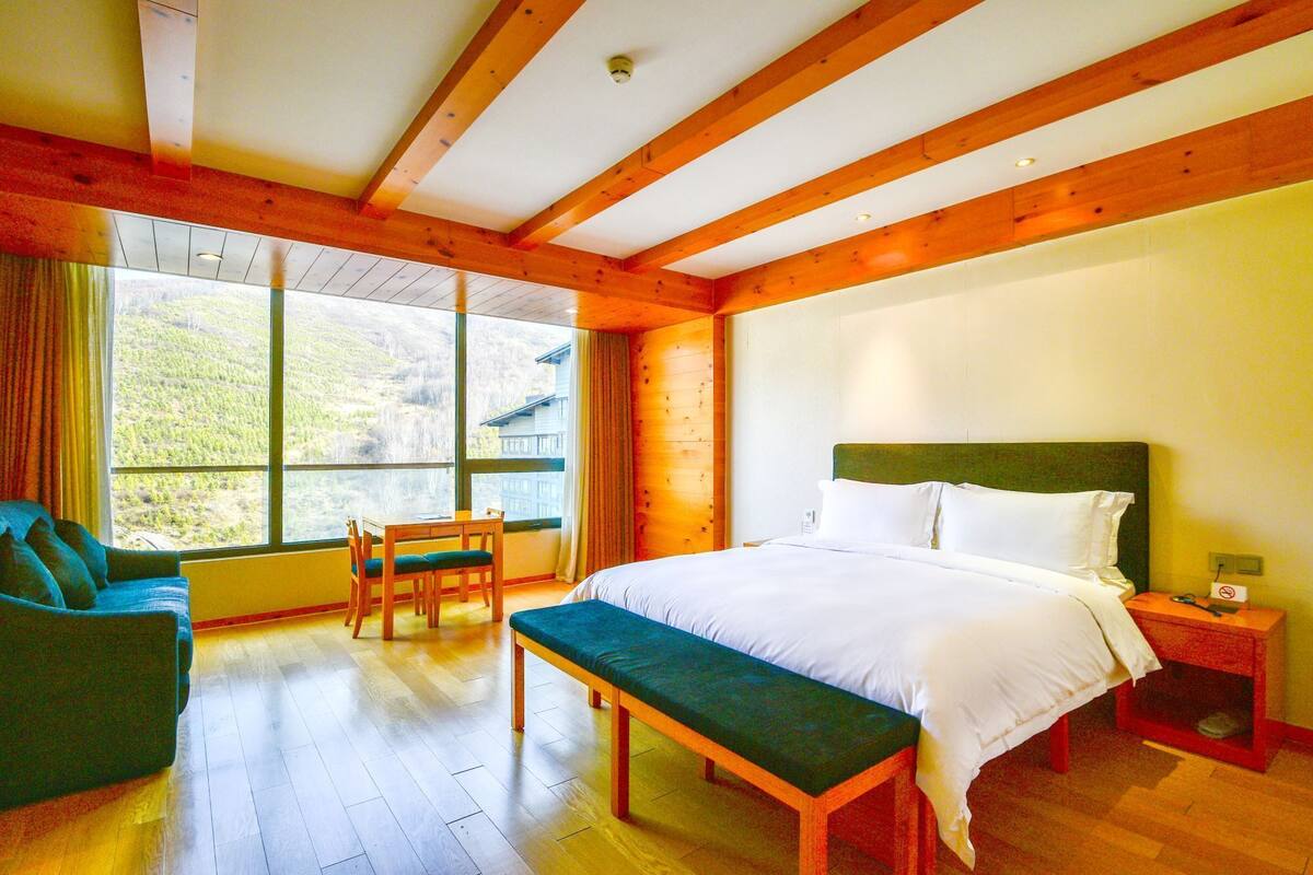 Un dormitorio con un ventanal grande, vigas de madera, una cama, un banco y una silla.