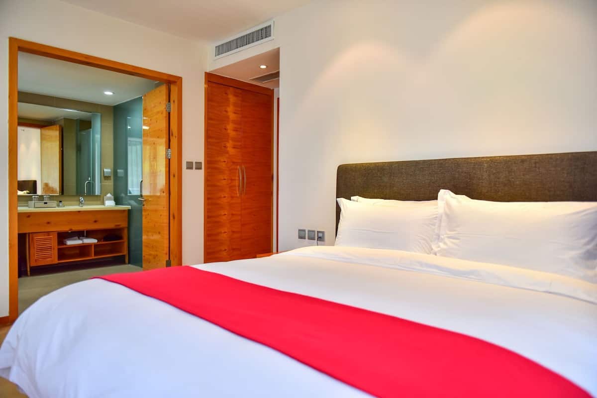 Habitación de hotel con una cama grande, un armario de madera, un baño con espejo y un detalle rojo en la colcha.