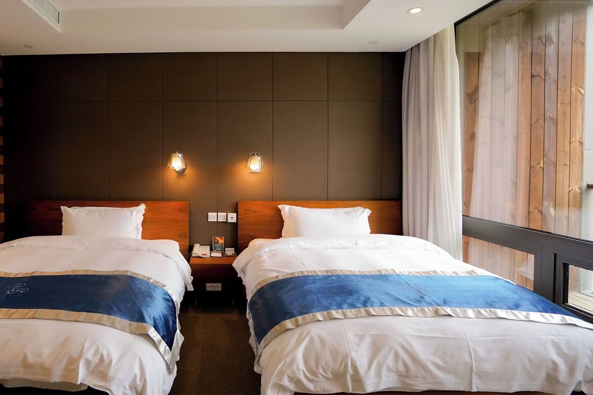 Habitación de hotel con dos camas, cabeceros de madera, un ventanal con cortinas e iluminación empotrada.