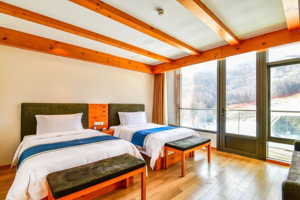 Habitación de hotel con dos camas, vigas de madera, un ventanal con vistas a un paisaje nevado y un banco.
