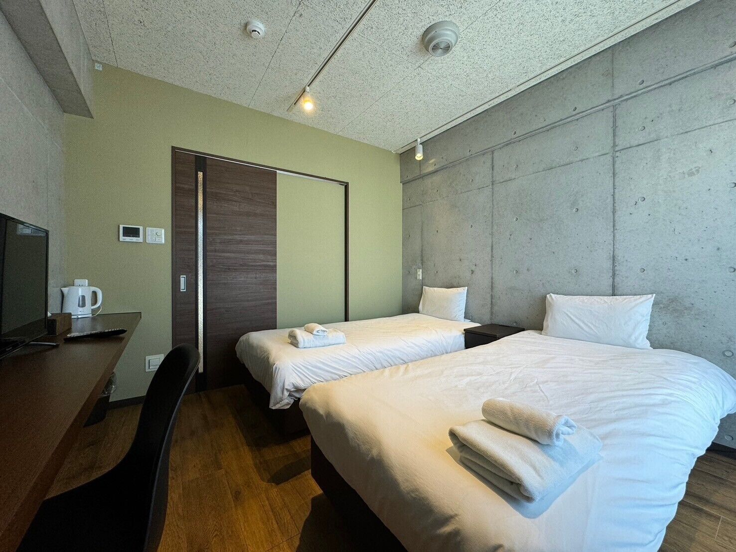 A hotel room with two beds, a desk with a computer and a coffee maker, a wardrobe, and a concrete accent wall.
