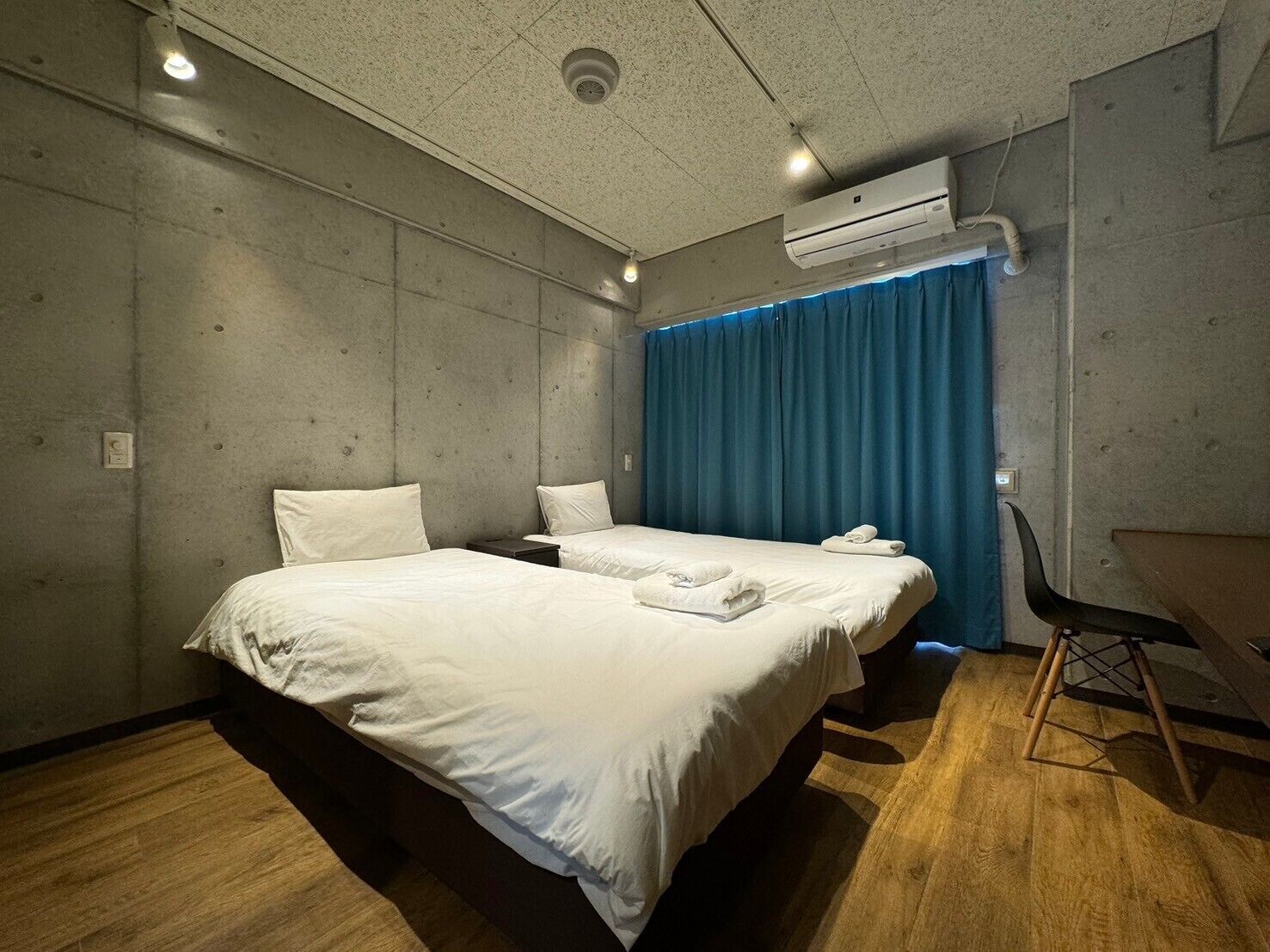 A hotel room with two beds, a desk, an air conditioning unit, and a blue curtain.