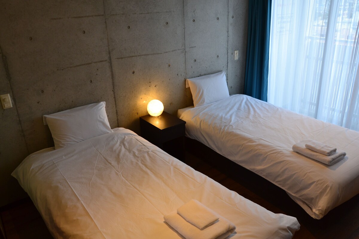 A hotel room with two single beds, a nightstand with a lamp, and a window with curtains.