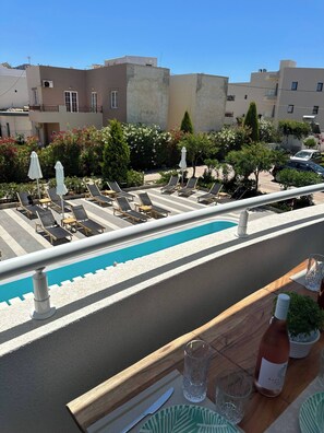 A rooftop pool area with lounge chairs and umbrellas, a wooden table with a bottle and glasses, and a view of surrounding buildings.