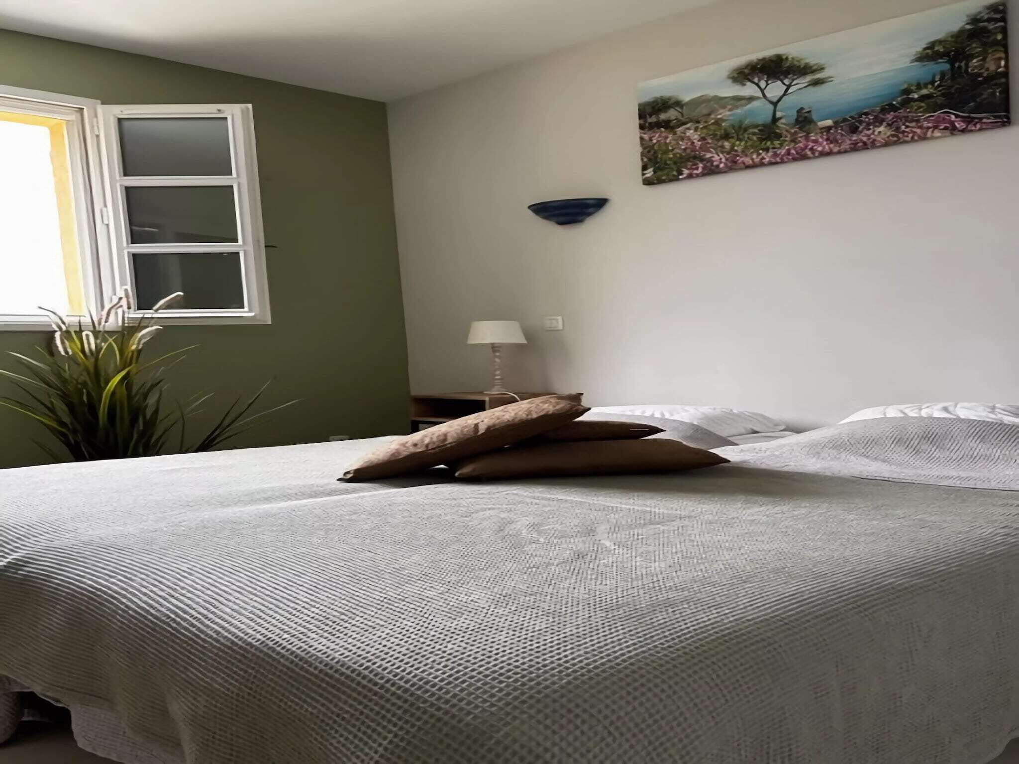 Un lit bien fait, avec des oreillers, une fenêtre avec des stores, une lampe fixée au mur et un tableau.