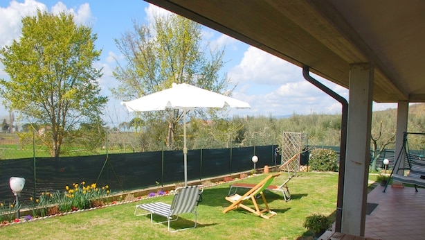 Een overdekt terras met een gazon, twee ligstoelen, een schommel en een houten tafel met een bloempot.