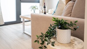 A modern living room with a beige sofa, a small round wooden table with a potted plant, and a wine bottle on the windowsill.