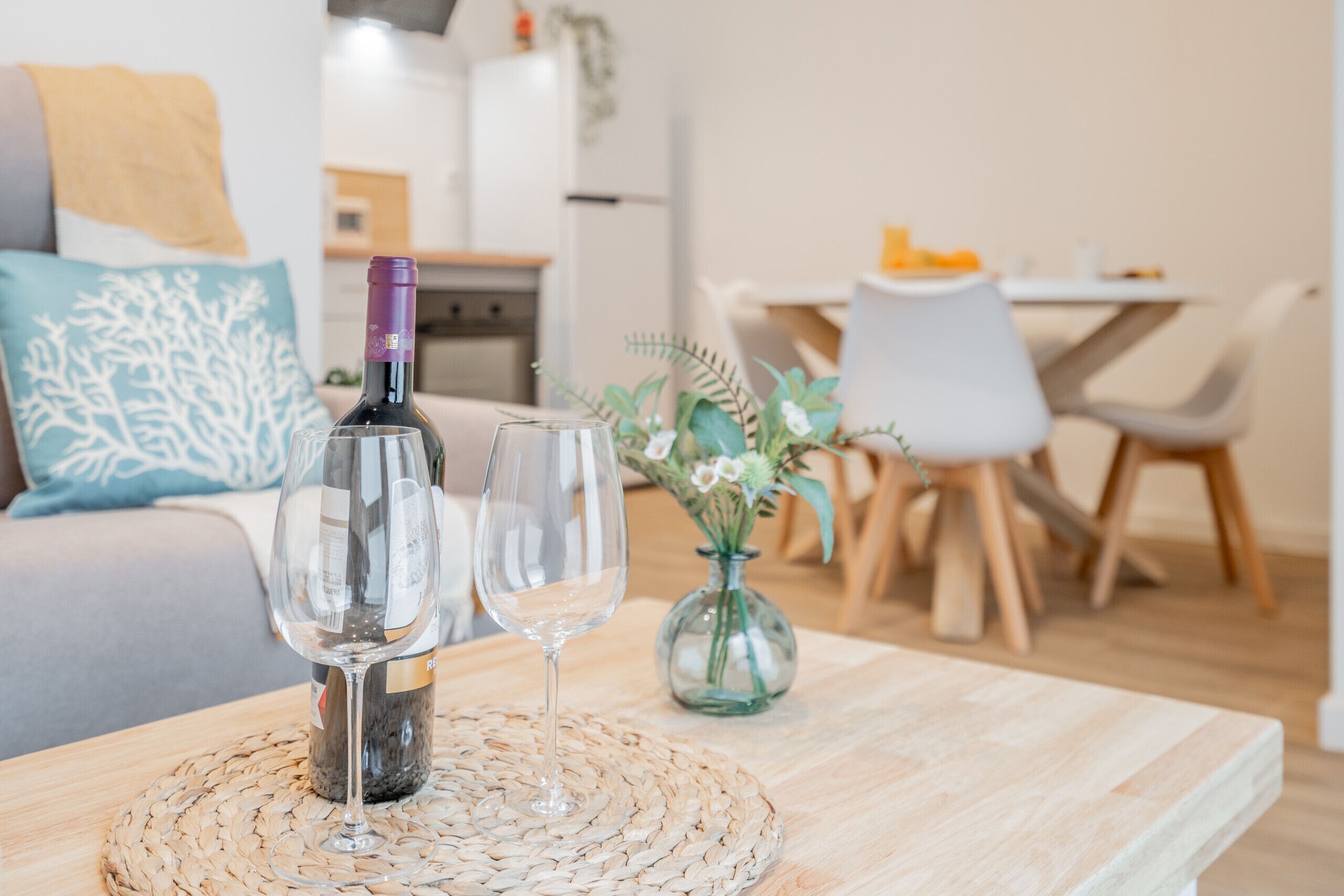 Een zithoek met een houten tafel, een fles wijn en twee glazen. Er staat een bank met een decoratief kussen en een vaas met bloemen op de achtergrond.