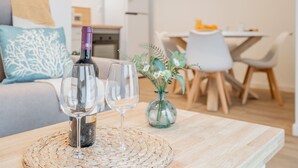 A living area with a wooden table, a bottle of wine, and two glasses. There is a sofa with a decorative pillow and a vase with flowers in the background.