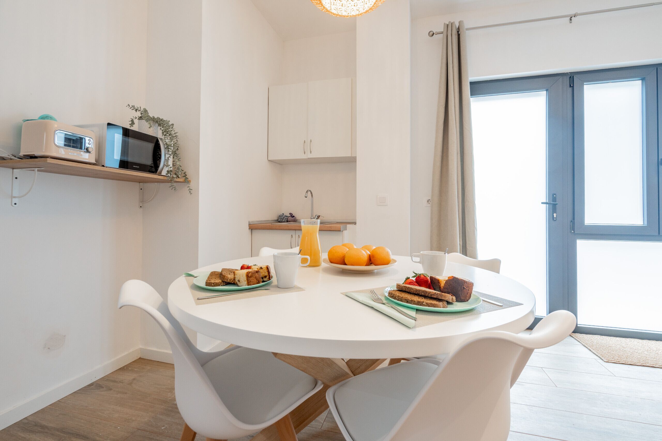 A modern kitchen with white cabinets, a microwave, and a toaster on a shelf. A dining table with chairs is set with breakfast items.