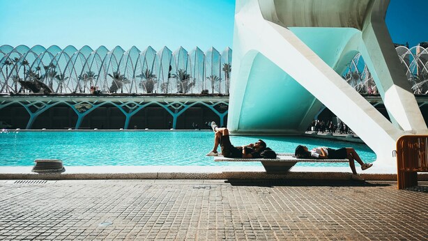 Un área de piscina con una estructura arquitectónica moderna de fondo, un camino de baldosas y algunas personas relajándose junto a la piscina.