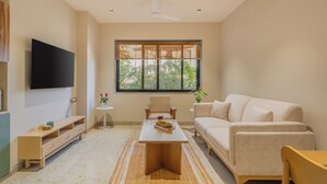 A modern living room with a flat-screen TV, a beige sofa, a wooden coffee table, and a large window with a view of greenery. - ELIVAAS Kamerios Verano 1 BHK with Pool (Marra)