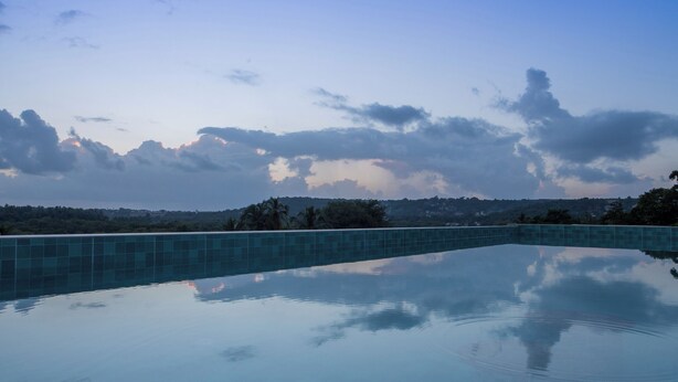 A clear pool with a tiled edge, surrounded by a low wall and a clear sky with clouds. - ELIVAAS Kamerios Verano 1 BHK with Pool (Marra)