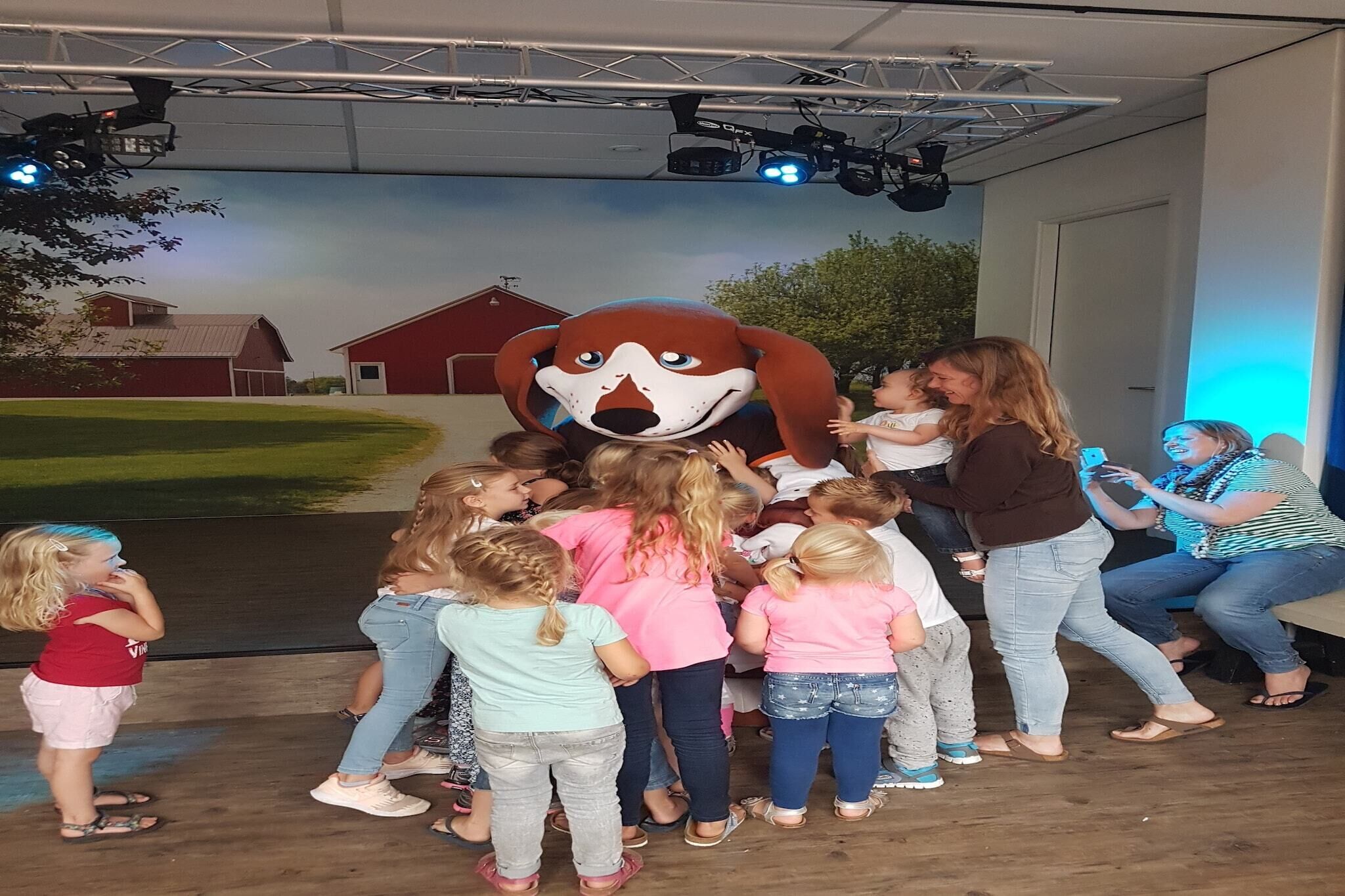 Un groupe d’enfants et d’adultes s’est rassemblé autour d’une grande statue représentant une mascotte, dans une pièce dont le mur du fond était décoré d’une fresque représentant une ferme et des granges.