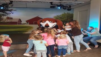 Un groupe d’enfants et d’adultes s’est rassemblé autour d’une grande statue représentant une mascotte, dans une pièce dont le mur du fond était décoré d’une fresque représentant une ferme et des granges.