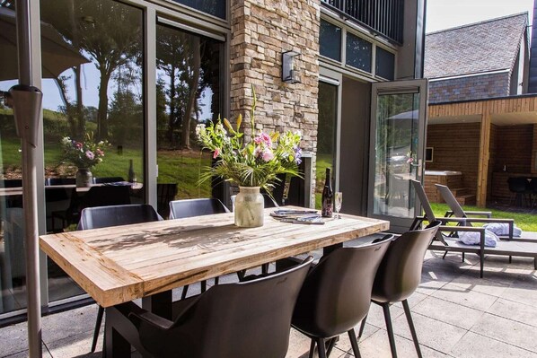 An outdoor dining area with a wooden table and chairs, a vase of flowers, and a stone wall.