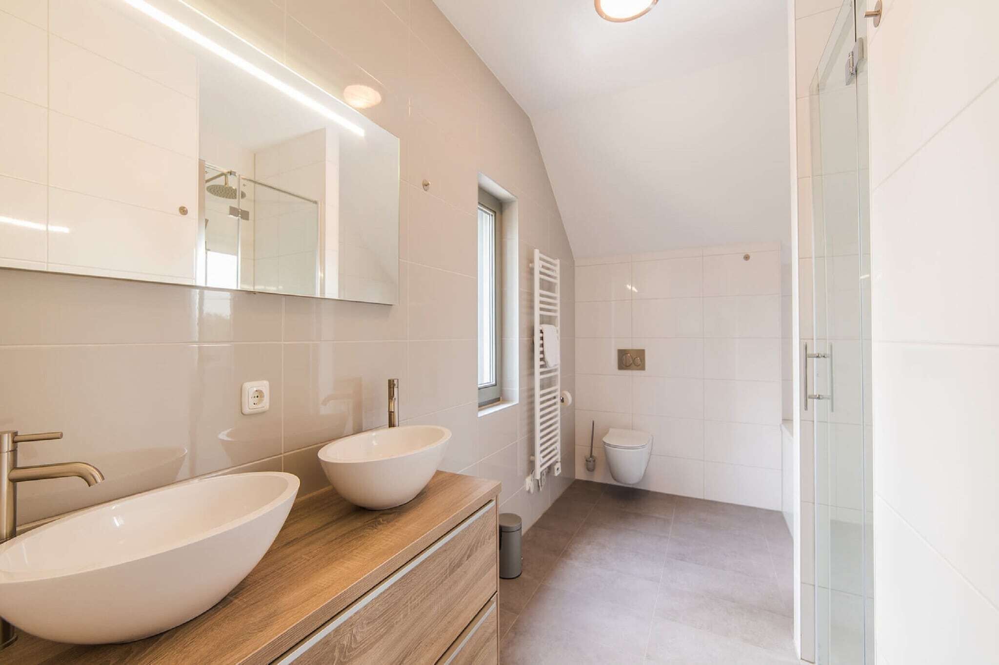 Een moderne badkamer met een grote spiegel, twee wasbakken, een houten wastafelmeubel, een douchecabine met glazen wanden en een wit toilet.