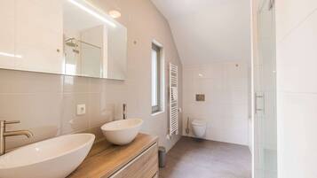 Een moderne badkamer met een grote spiegel, twee wasbakken, een houten wastafelmeubel, een douchecabine met glazen wanden en een wit toilet.
