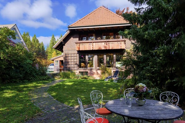 A wooden house with a balcony, surrounded by greenery and a paved path leading to a round table with chairs. - Altes Forsthaus Kniebis | almost secluded location | historic | dog | barbecue | WLAN (Freudenstadt)