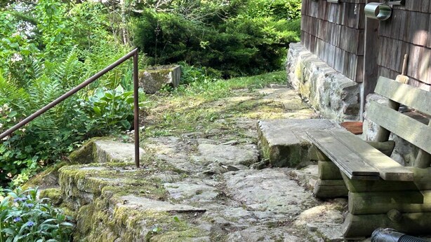 Una escalera de piedra conduce a un banco de madera, rodeado de follaje y un edificio de madera.