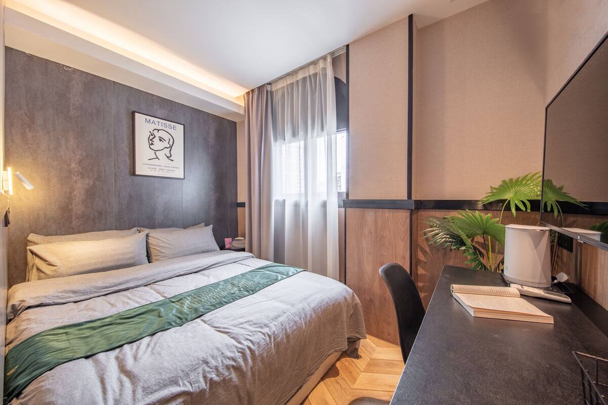 A hotel room with a bed, a desk with a lamp, a plant, and a wall-mounted artwork.