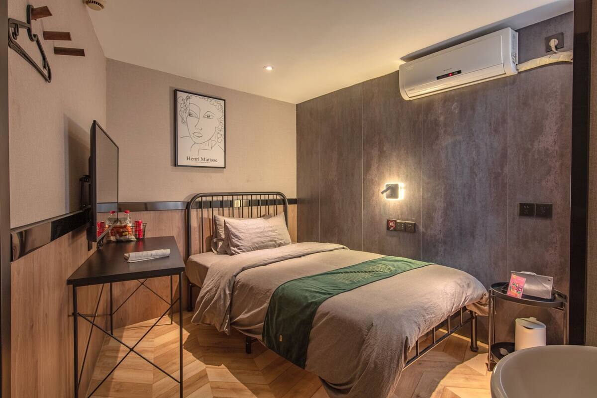 A hotel room with a bed, a desk, a chair, a wall-mounted air conditioner, and a framed artwork.