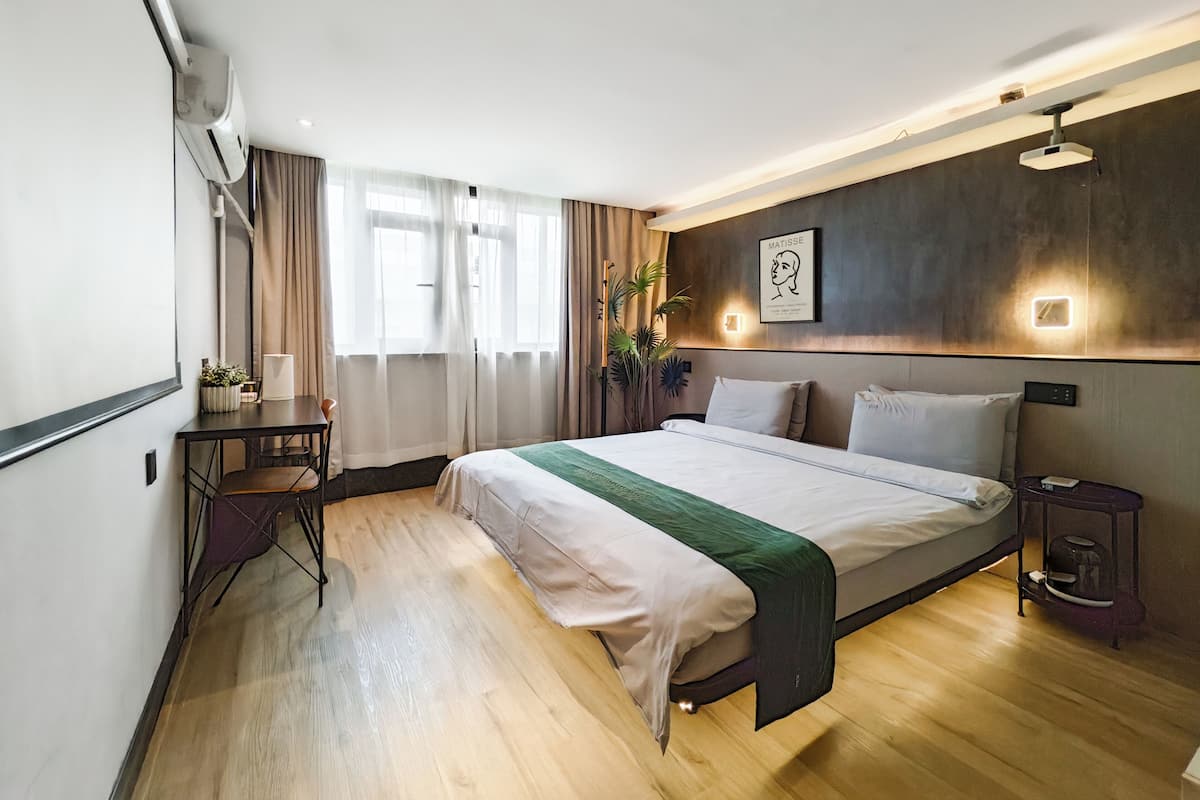 A modern hotel room with a large bed, a desk, a chair, and a bedside table with a lamp.
