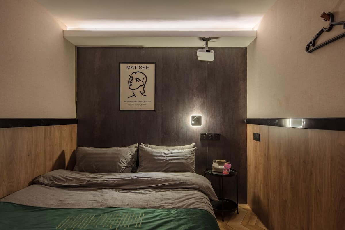 A modern bedroom with a wooden headboard, a bed with a green bedspread, a wall-mounted light, and a framed artwork of a woman's profile.