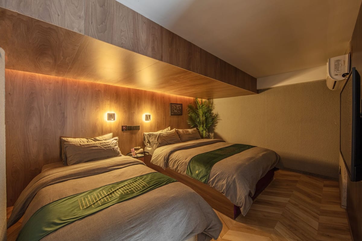 A hotel room with two beds, wooden paneling, wall-mounted lights, a plant, and a small table.