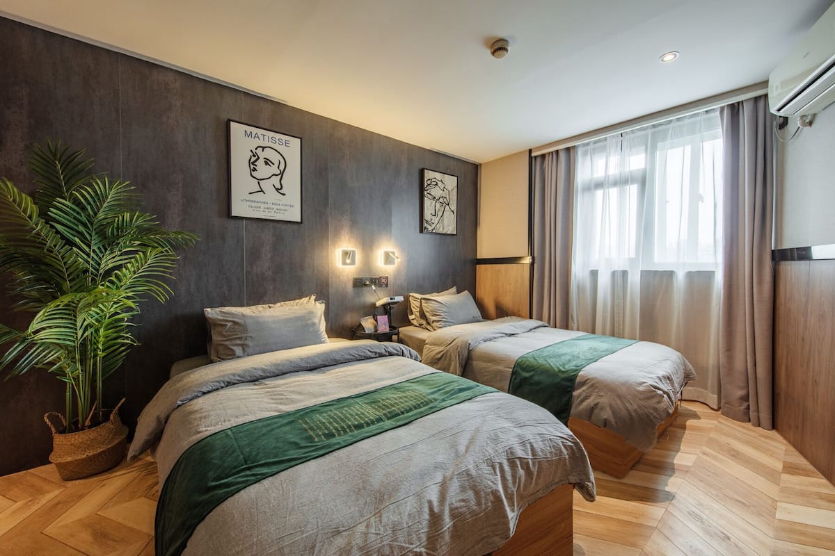 A modern hotel room with two beds, a large potted plant, artwork on the walls, and a large window with curtains.