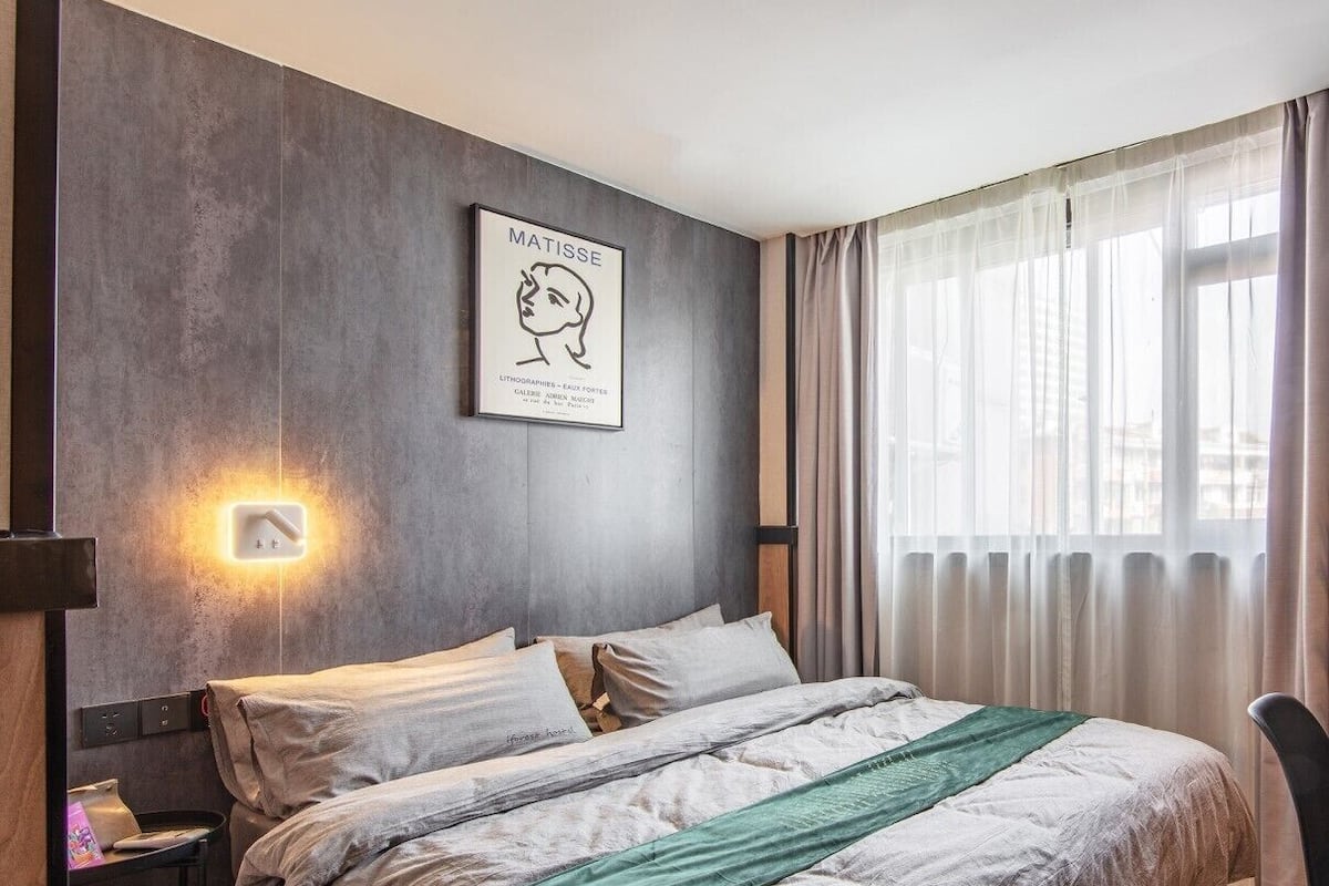 A modern bedroom with a bed, a nightstand, a wall-mounted light, and a window with curtains.