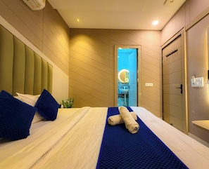 Quarto de hotel com uma cama, um tapete azul texturizado, uma toalha, uma mesinha de cabeceira, um espelho e um banheiro visível através de uma porta aberta.