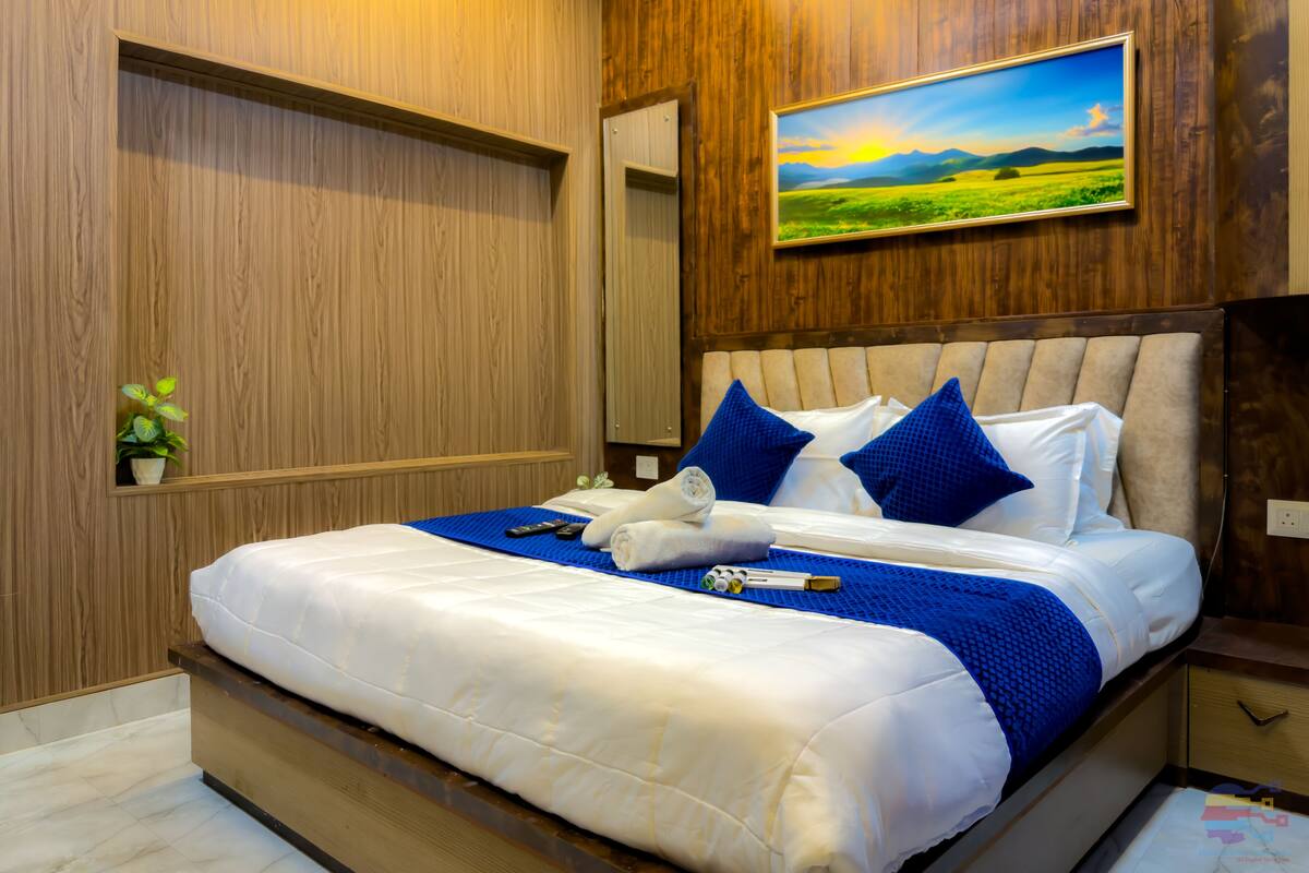 A bedroom with a large bed, wooden panel walls, a painting of a landscape, and a small plant.