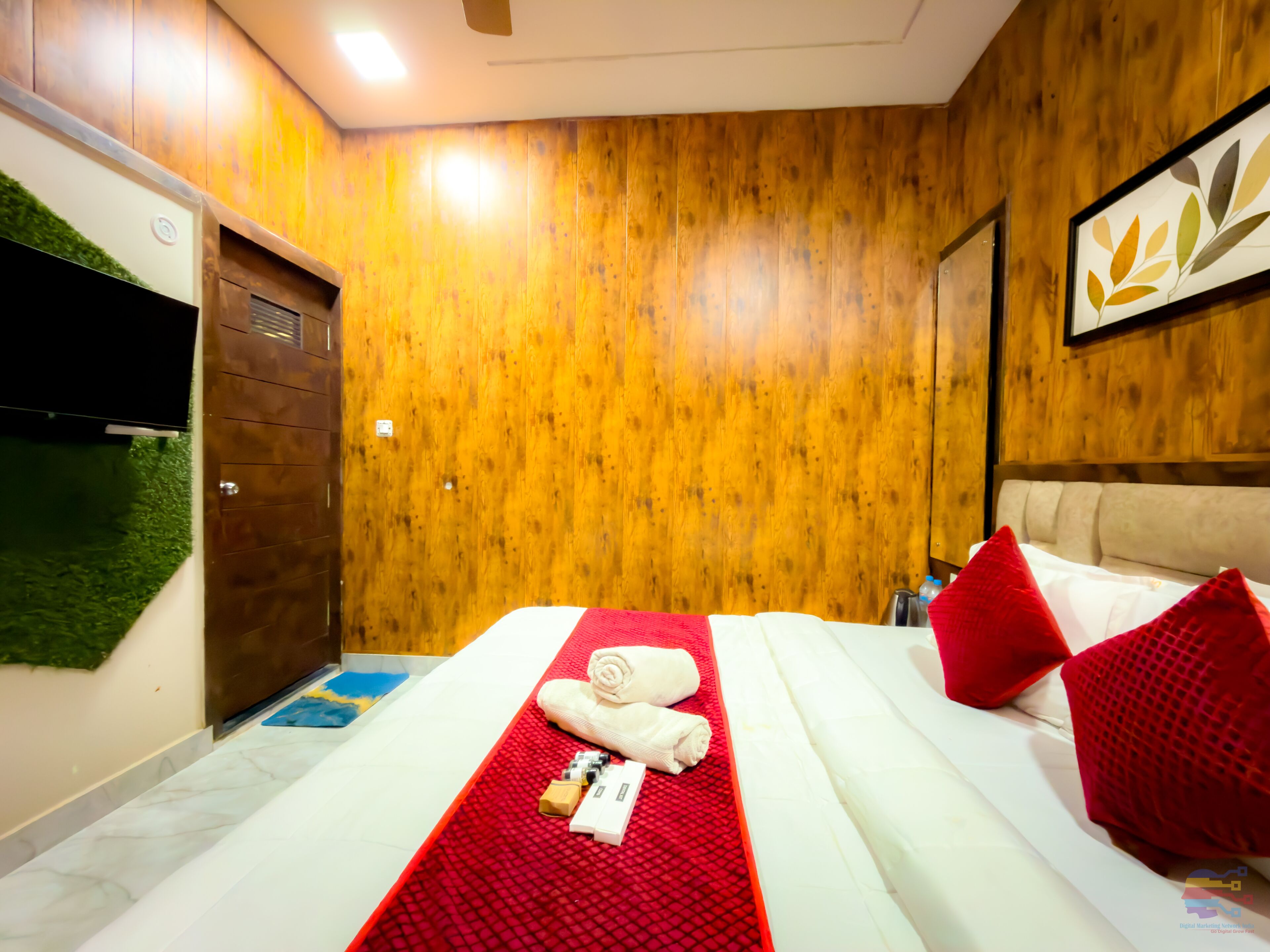 A hotel room with wooden walls, a bed with a red bedspread, a white headboard, and a painting of leaves.