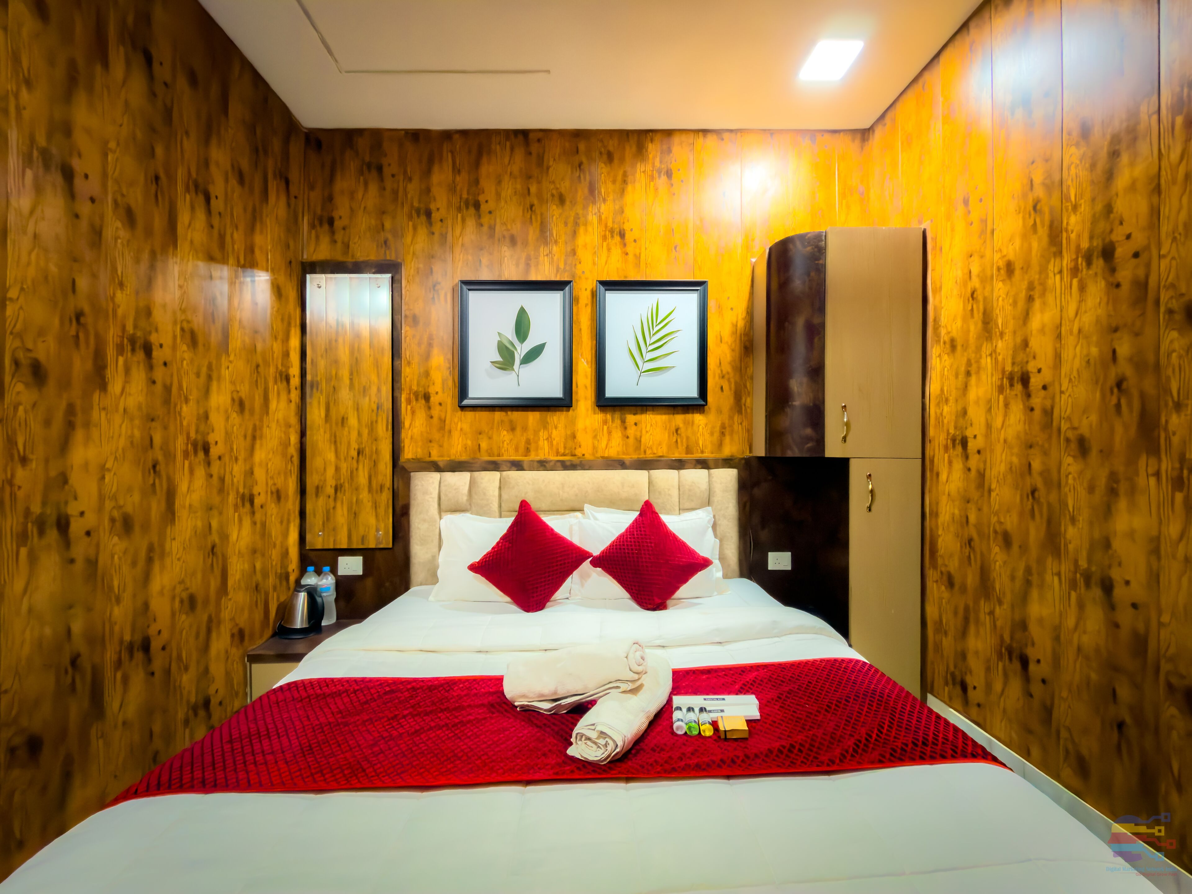 Habitación de hotel con paredes de madera, cama con ropa de cama roja y blanca, dos adornos con hojas enmarcados, una minibar y un armario.