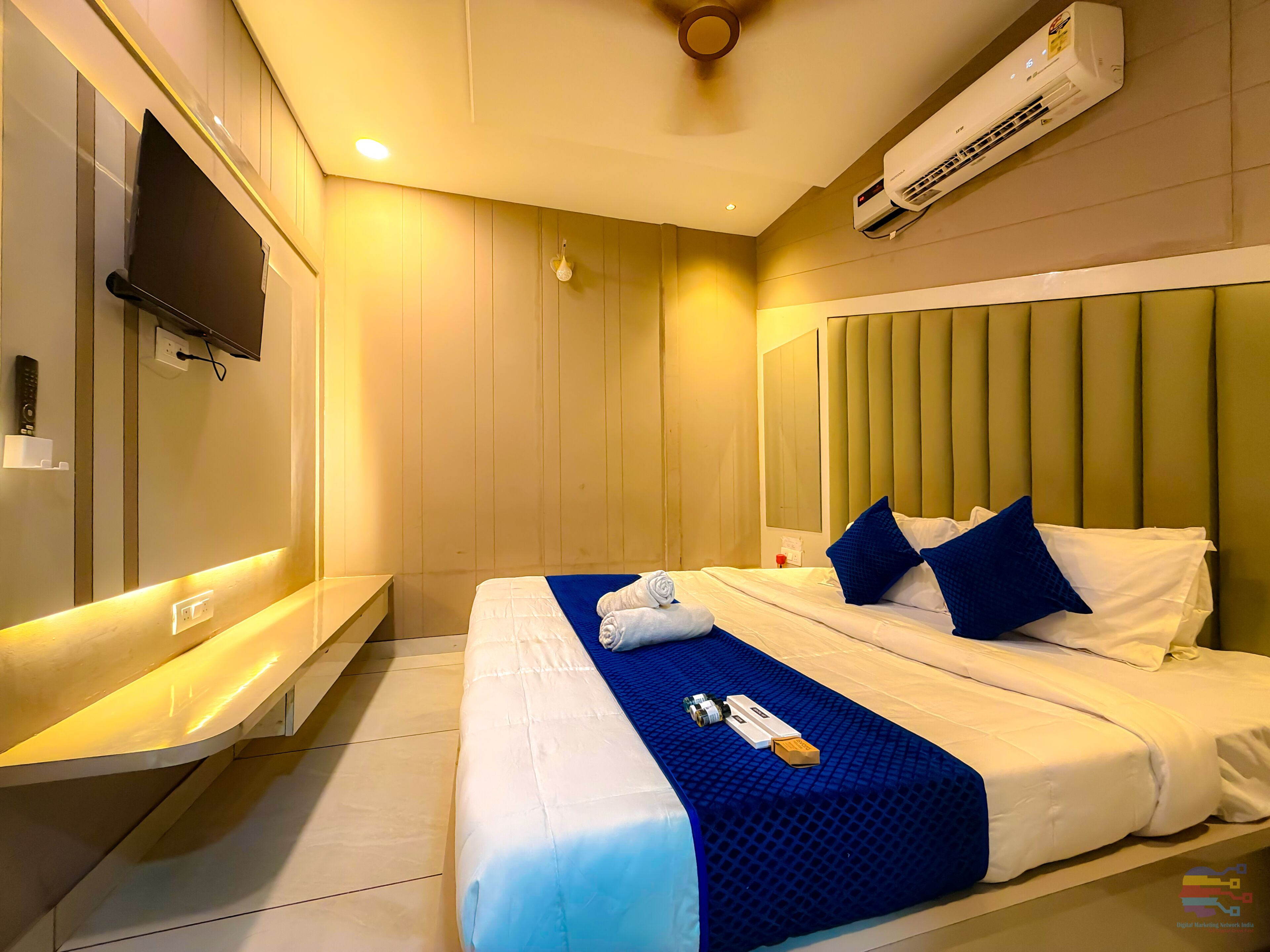A hotel room with a bed, bedside table, TV, air conditioning unit, and a wall-mounted light fixture.