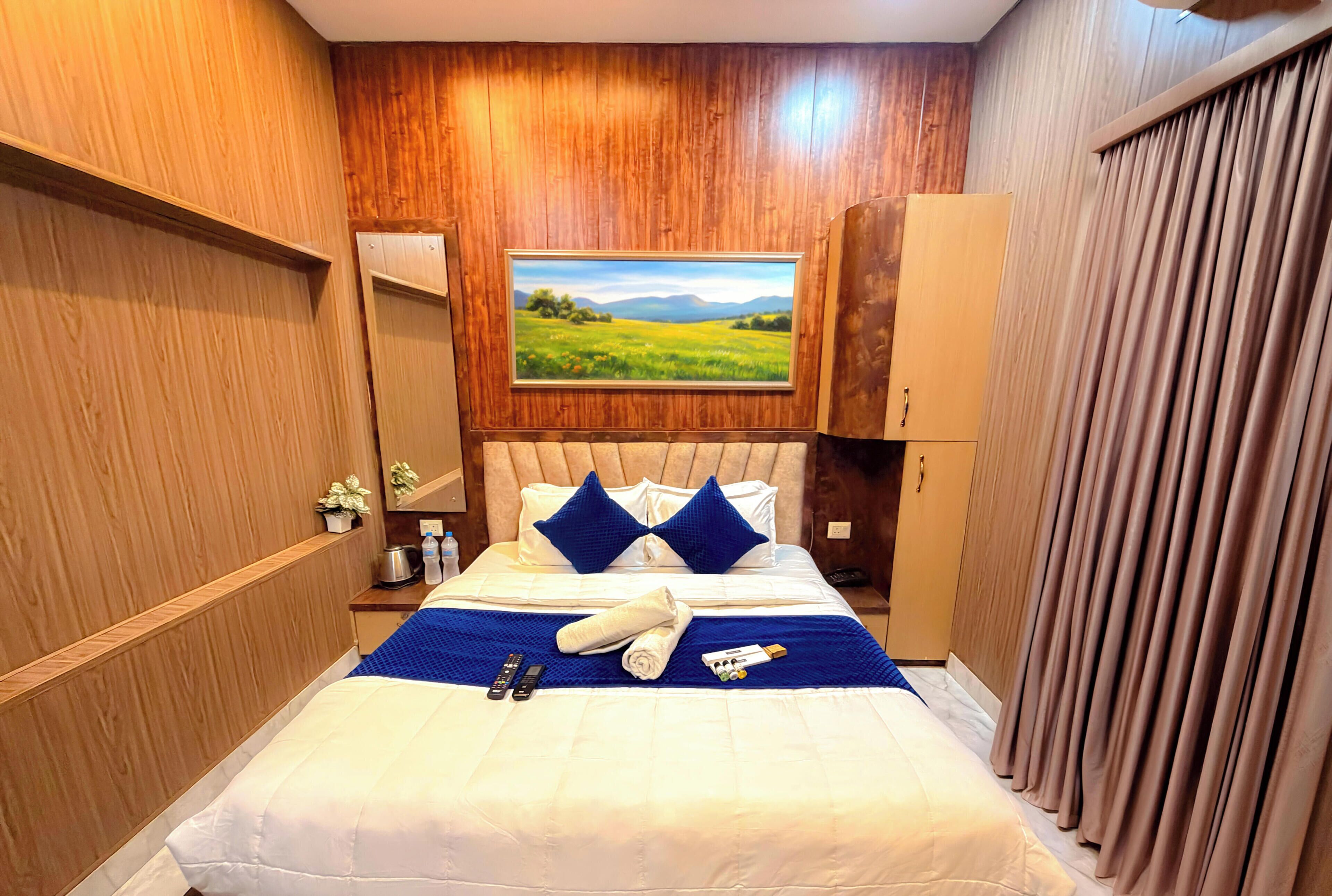 A neatly made bed with a blue blanket, pillows, and a rolled towel. There is a painting of a landscape on the wall above the bed.