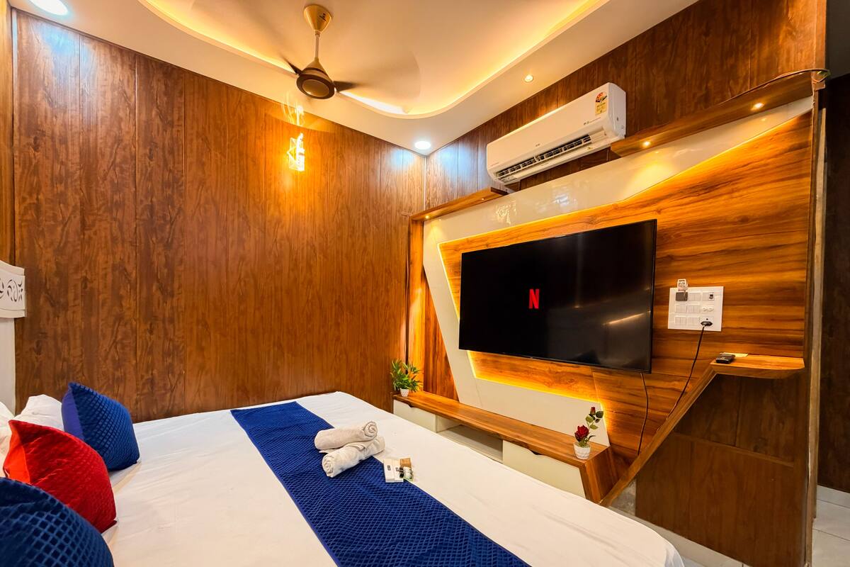 A modern hotel room with a large bed, a flat-screen TV mounted on a wooden wall, an air conditioning unit, and a ceiling fan.