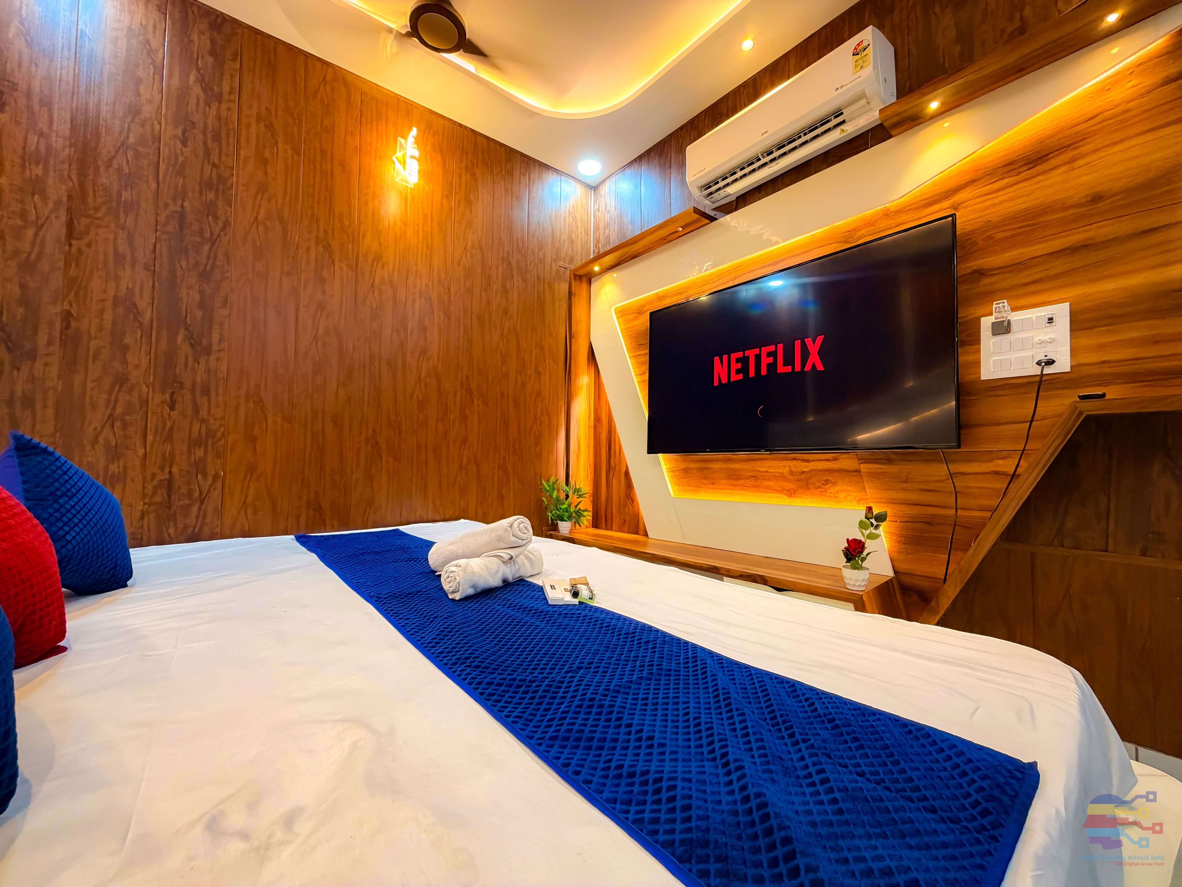 A hotel room with a large bed, a flat-screen TV, wooden paneling, and an air conditioning unit.