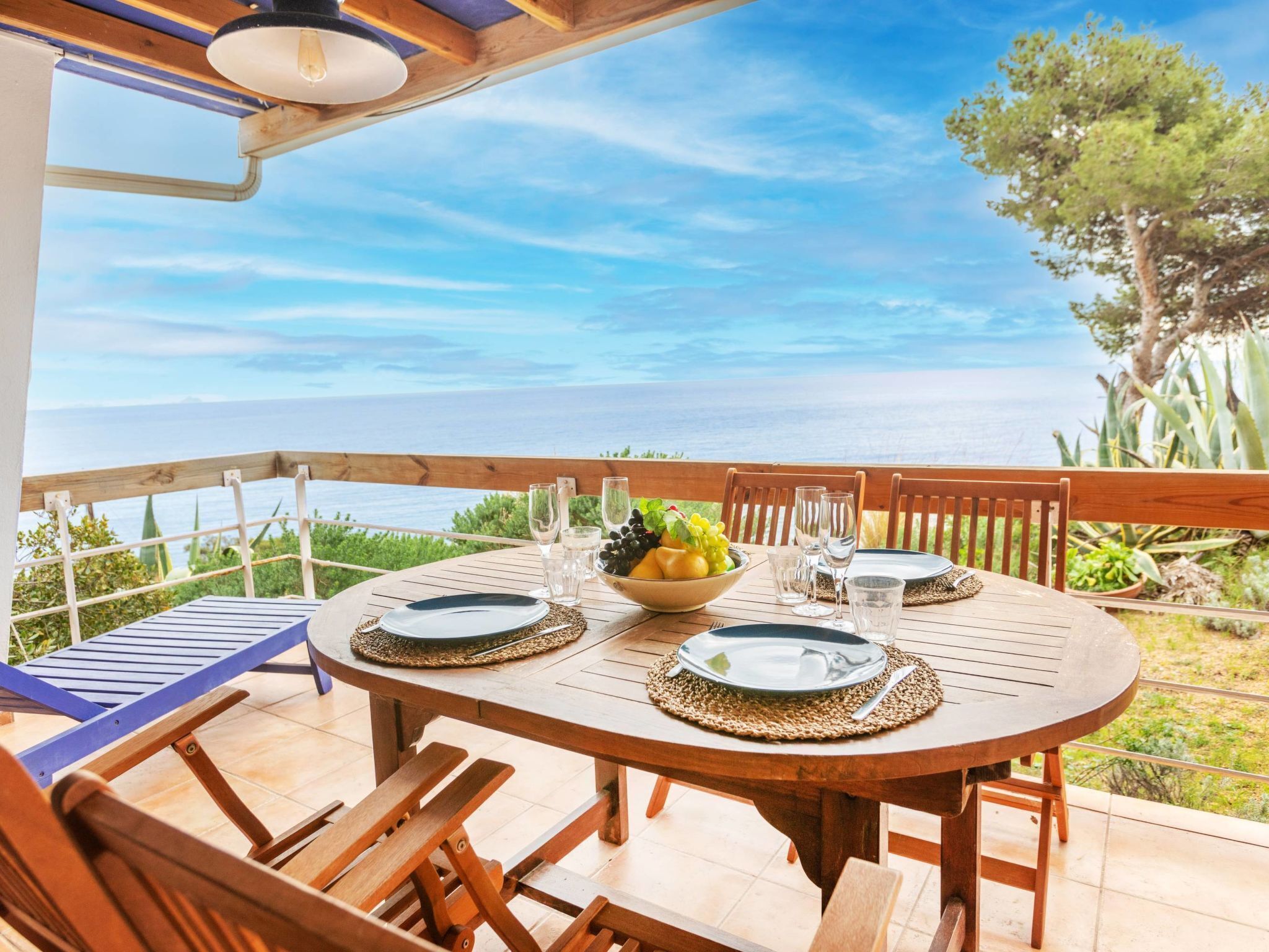 A wooden table set for two with a bowl of fruit, glasses, and plates on a balcony overlooking a body of water.