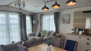 A modern living room with a sofa, a wooden dining table, and a kitchen area with a kettle and tea set. - Luxury holiday Home close to Padstow and beautiful beaches  (Saint Merryn)