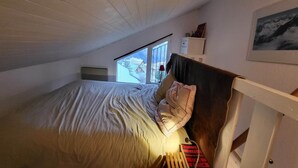 Ein Schlafzimmer mit schrägem Dach, einem Bett mit Kopfteil, einem Fenster mit Blick auf eine verschneite Landschaft und einem kleinen Regal mit Gegenständen an der Wand.